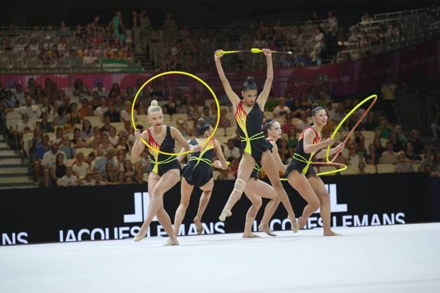 A group of five gymnasts in black leotards with red-and-yellow flame appliqués performs a synchronized group routine with yellow hoops and clubs at the Festhalle in Frankfurt. The athletes are arranged in a dynamic formation, while the audience fills the packed hall. Group routine with hoops and clubs at the 2026 Rhythmic Gymnastics World Championships Copyright: xspeedshotx