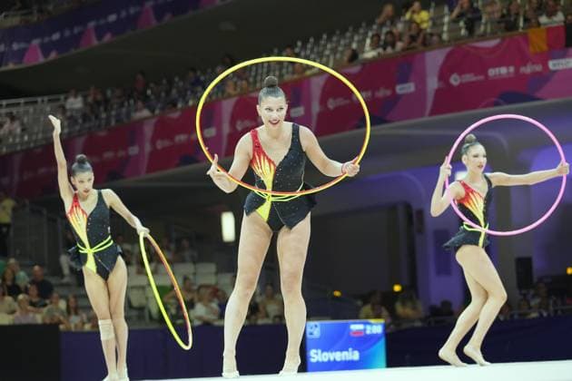 Three gymnasts from the Slovenian ensemble perform their group routine with hoops at the Festhalle in Frankfurt. The scoreboard in the background displays Slovenia, confirming their participation in the 2026 World Championships. Slovenian ensemble performing with hoops at the Rhythmic Gymnastics World Championships in Frankfurt Copyright: xspeedshotx