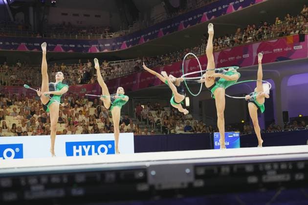 The Estonian group team performs a synchronized group routine with hoops and clubs at the 2026 Rhythmic Gymnastics World Championships in Frankfurt’s Festhalle. Four gymnasts in green and black leotards simultaneously perform high leg extensions, while holding the apparatus in the air. Estonia’s group team with hoops and clubs at the Rhythmic Gymnastics World Championships Copyright: xspeedshotx