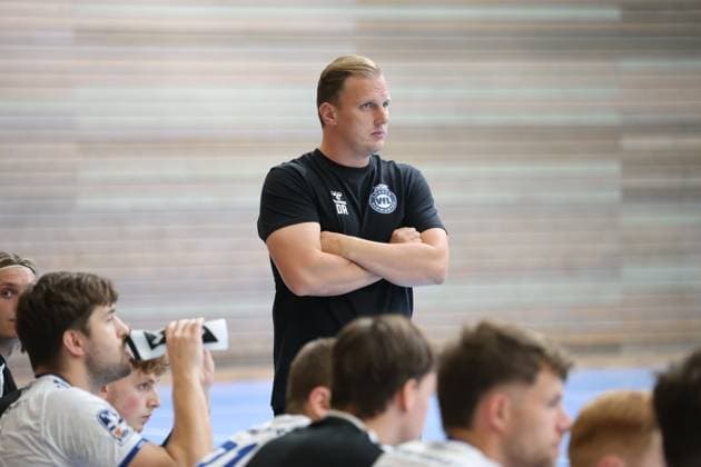 VfL Lübeck Schwartau coach David Röhrig stands on the sideline with his arms crossed, focused, watching the game at the Hansehalle in Lübeck. In the background, several players are sitting on the bench, including Guy Resch Ermann. David Röhrig watches the action from the sideline. Copyright: xspeedshotx