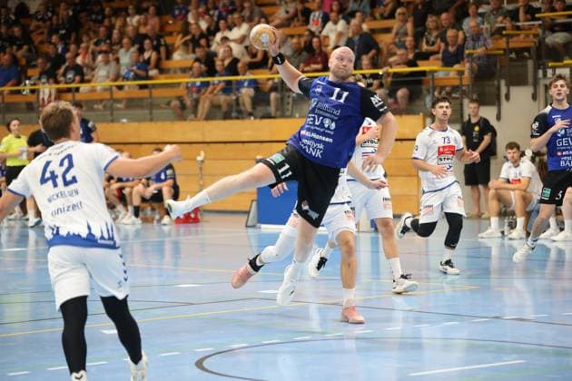 Kristoffer Jessen, 11, of Grindstedt GIF, jumps and holds the ball above his head, ready to shoot, while Lukas Kingsley Emdorf 73 of VfL Lübeck Schwartau and Einar Nickelsen 42 of VfL Lübeck Schwartau watch the action. In the background, other players from both teams, as well as a packed stands, watch the action unfold. Kristoffer Jessen 11 of Grindstedt GIF takes a jump shot at the Hansehalle. Copyright: xspeedshotx
