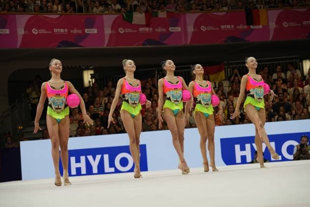 Five gymnasts from a group perform their synchronized ball routine on the competition mat at the Festhalle in Frankfurt. The athletes are wearing identical pink, yellow, and turquoise competition leotards, and each is holding a pink ball. Group ball routine at the Rhythmic Gymnastics World Championships in Frankfurt Copyright: xspeedshotx