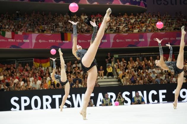 Three gymnasts from a group perform a synchronized ball routine at the Festhalle in Frankfurt, throwing the pink balls high into the air. The athletes perform expressive stretch-and-spread jumps in front of a sold-out audience. Group routine with ball at the 2026 Rhythmic Gymnastics World Championships in Frankfurt Copyright: xspeedshotx