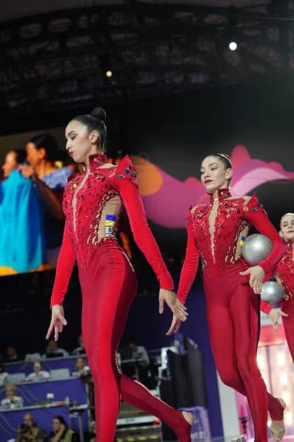 Three gymnasts from the Ukrainian group team, wearing red competition leotards, perform their routine at the Festhalle in Frankfurt; two of them are holding silver balls. In the background, other athletes and the colorful World Championships decorations are visible. The Ukrainian group during their group routine – 2026 Rhythmic Gymnastics World Championships in Frankfurt Copyright: xspeedshotx