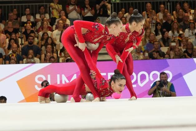 Five gymnasts in red leotards are performing a synchronized group routine with balls in Frankfurt’s Festhalle. One gymnast is lying on the floor, while her teammates lean over her and control the silver and pink balls. Group gymnastics team performing a synchronized ball routine at the 2026 World Championships Copyright: xspeedshotx