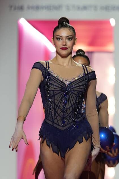 A gymnast from Team Spain, wearing a dark blue competition leotard studded with rhinestones, strides into the Festhalle Frankfurt during the entrance parade. In the background, another gymnast can be seen holding a blue ball. Team Spain at the entrance parade for the 2026 Rhythmic Gymnastics World Championships Copyright: xspeedshotx