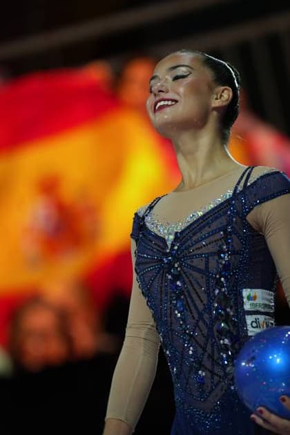 A gymnast from Spain is holding a blue ball and smiling with her eyes closed; with the Spanish flag visible in the background. The photo was taken at the 2026 Rhythmic Gymnastics World Championships in the Festhalle in Frankfurt. Spanish gymnast with a ball and a beaming smile at the Rhythmic Gymnastics World Championships Copyright: xspeedshotx