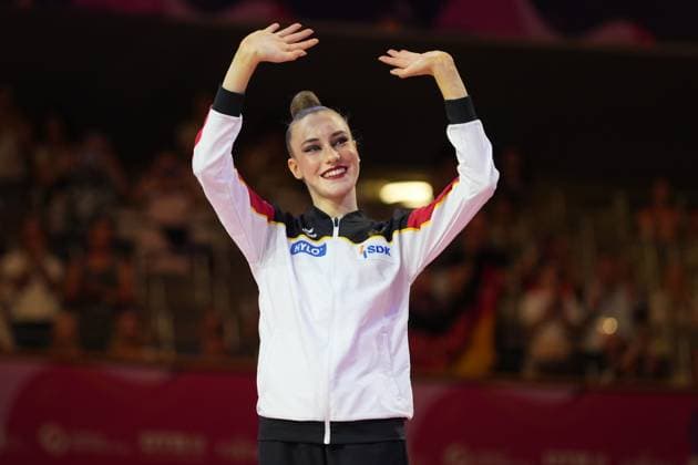 A gymnast in the German national uniform raises her arms high with a beaming smile and celebrates in front of the audience at the Festhalle in Frankfurt. She is wearing the German team’s white, black, red, and gold training jacket featuring the sponsors HYLO and SDK. A German gymnast cheers at the Rhythmic Gymnastics World Championships in Frankfurt. Copyright: xspeedshotx