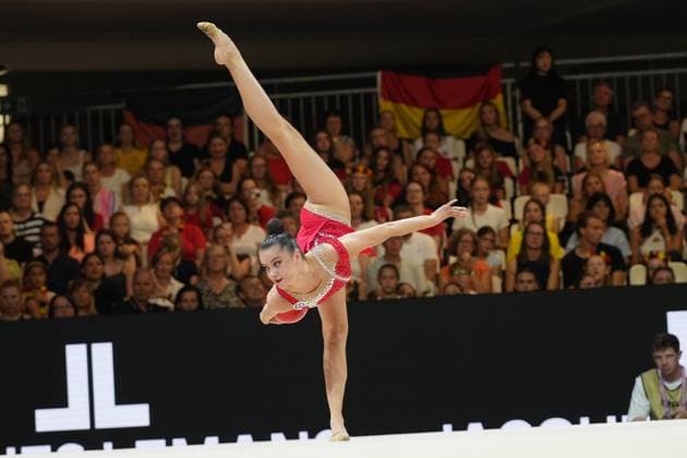 A gymnast in a red competition leotard performs a balance element with her leg extended straight upward. In the background, numerous spectators watch the routine at the Festhalle in Frankfurt; a German flag is visible. Gymnast performing a balance element with her leg extended straight upward at the Rhythmic Gymnastics World Championships Copyright: xspeedshotx