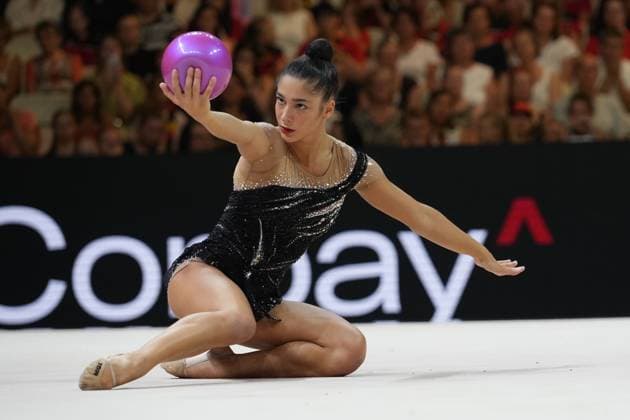 The Italian gymnast is performing a floor routine with a ball at the Festhalle in Frankfurt, sitting low to the floor and extending the ball upward with her arm outstretched. In the background, the packed audience and an advertising banner are visible. Italian gymnast with a ball at the Rhythmic Gymnastics World Championships in Frankfurt Copyright: xspeedshotx