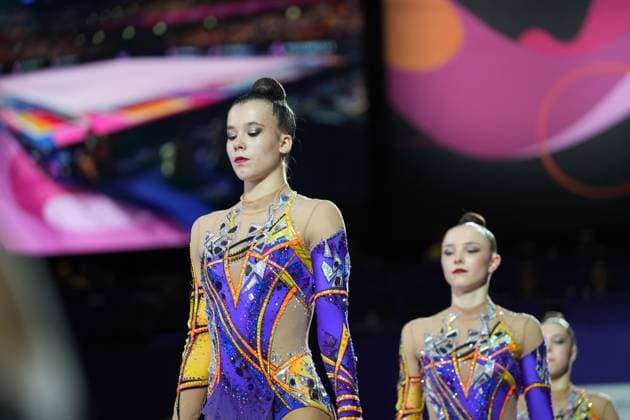 Three gymnasts from Germany, wearing identical purple, yellow, and orange competition leotards, stand intently in the Festhalle Frankfurt. The athletes appear focused and absorbed in their thoughts before their performance at the 2026 Rhythmic Gymnastics World Championships – Gymnasts before their performance at the Festhalle in Frankfurt Copyright: xspeedshotx