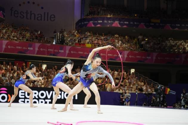 Three gymnasts from Team Japan are performing a synchronized group routine with hoops and clubs at the Festhalle in Frankfurt, while the audience fills the packed hall. In the foreground, a pink rope can be seen on the floor, indicating another piece of equipment used in the routine. Team Japan’s group routine with hoops and clubs at the Rhythmic Gymnastics World Championships in Frankfurt Copyright: xspeedshotx