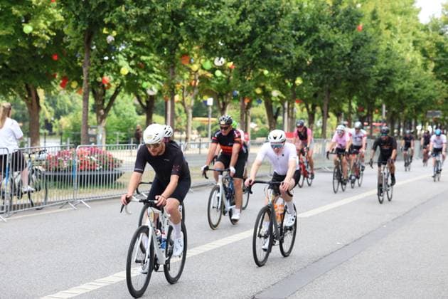 A group of cyclists rides down a closed-off street in Hamburg, lined with trees and colorful decorations. In the foreground, a rider in a black jersey and white helmet leads the pack of cyclists in the peloton at the Cyclassics Hamburg. Copyright: xspeedshotx xSarahxWolfx