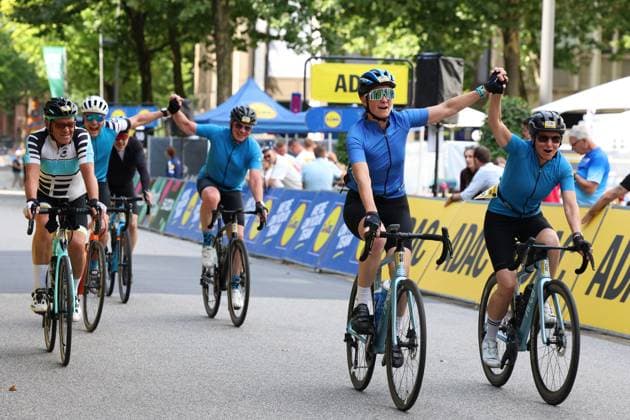 Several participants in the Cyclassics Hamburg cross the finish line at Rathausmarkt, cheering and raising their arms in the air. In the background, ADAC and Lidl advertising banners and a blue tent are visible. Cheering amateur cyclists cross the finish line at the Cyclassics Hamburg. Copyright: xspeedshotx xSarahxWolfx