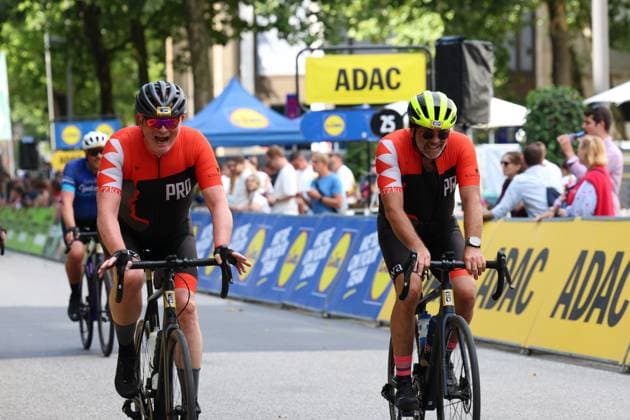 Two riders in red and black jerseys cross the finish line together, laughing, at Hamburg’s Rathausmarkt. ADAC and Lidl advertising banners and spectators are visible in the background. Two amateur riders crossing the finish line at the Cyclassics Hamburg Copyright: xspeedshotx xSarahxWolfx