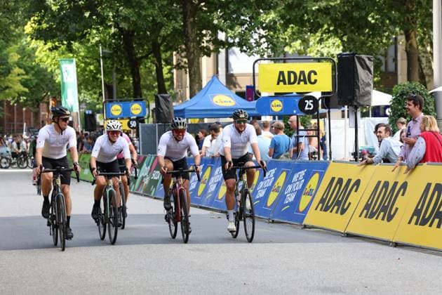 Four cyclists in white jerseys cross the finish line together at Hamburg’s Rathausmarkt, flanked by ADAC and Lidl advertising banners. In the background, numerous spectators watch the race. Cyclists pass the finish line at the Cyclassics Hamburg. Copyright: xspeedshotx xSarahxWolfx