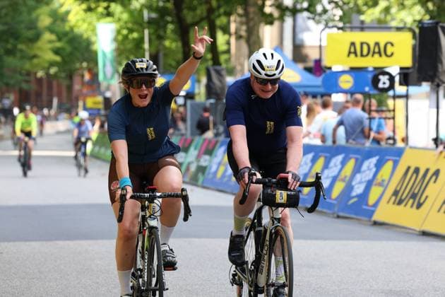 Two participants in dark blue jerseys cross the finish line together at Hamburg’s Rathausmarkt; one of them raises his arm in celebration. In the background, ADAC and Lidl advertising banners can be seen, as well as other cyclists. Two amateur cyclists celebrate as they cross the finish line at the Cyclassics Hamburg. Copyright: xspeedshotx xSarahxWolfx
