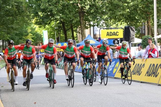 A group of cyclists wearing red-and-green GlückStolz jerseys crosses the finish line of the Cyclassics Hamburg at Rathausmarkt together and celebrates with arms outstretched. ADAC advertising banners and spectators are visible in the background. A cheering group at the finish of the Cyclassics Hamburg at Rathausmarkt Copyright: xspeedshotx xSarahxWolfx