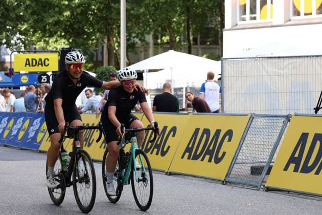 Two cyclists in black SKS jerseys are riding side by side at Hamburg’s Rathausmarkt; one has an arm around the other—both are smiling at the camera. ADAC advertising banners and spectators can be seen in the background. Two cyclists in black jerseys at the Cyclassics Hamburg race at Rathausmarkt Copyright: xspeedshotx xSarahxWolfx