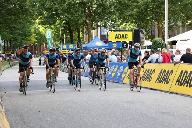 A group of amateur cyclists in turquoise and dark blue jerseys cross the finish line cheering at Hamburg’s Rathausmarkt during the Cyclassics. Several riders raise their arms in the air, with ADAC and Lidl advertising banners and spectators visible in the background. Amateur cyclists celebrate crossing the finish line at the Cyclassics in Hamburg Copyright: xspeedshotx xSarahxWolfx