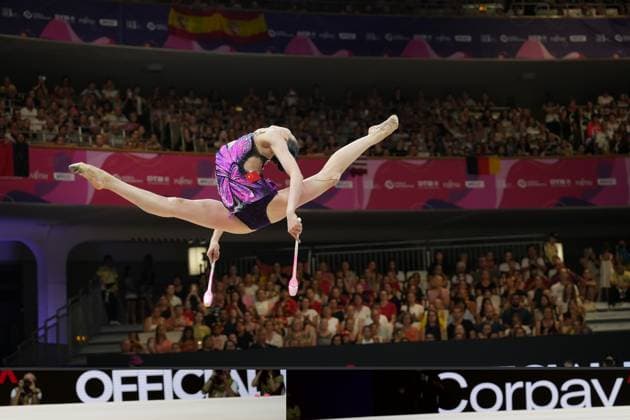 A gymnast from China performs a split jump with two clubs at the Rhythmic Gymnastics World Championships in Frankfurt’s Festhalle. The packed audience watches the routine in the background. Chinese gymnast with clubs performing a jump at the Rhythmic Gymnastics World Championships in Frankfurt Copyright: xspeedshotx