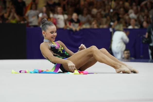 Sofiia Ilterikova sits smiling on the competition floor of the Festhalle in Frankfurt, striking her final pose after her routine with colorful ribbons. The colorful ribbons in yellow, pink, blue, and purple are spread out decoratively around her. Sofiia Ilterikova strikes her final pose with ribbons at the Rhythmic Gymnastics World Championships Copyright: xspeedshotx