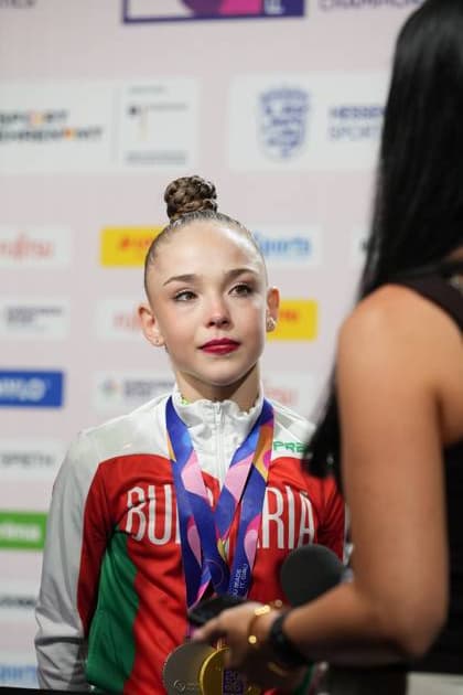 A gymnast from Bulgaria stands in front of a press barrier with several medals around her neck, giving an interview. In the background are sponsor logos for the 2026 Rhythmic Gymnastics World Championships at the Festhalle in Frankfurt. Bulgarian gymnast with a gold medal during an interview in Frankfurt Copyright: xspeedshotx