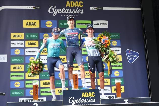 Paul Magnier 21 of Soudal Quick-Step stands on the top step of the podium at the ADAC Cyclassics Hamburg at Rathausmarkt, flanked by Laurence Pithie 31 of Red Bull – Bora – hansgrohe in third place and another Astana rider in second place. All three riders are holding bouquets of flowers; Erdinger beer glasses are on the podium. Paul Magnier (No. 21) wins the ADAC Cyclassics Hamburg – award ceremony on the podium Copyright: xspeedshotx xSarahxWolfx