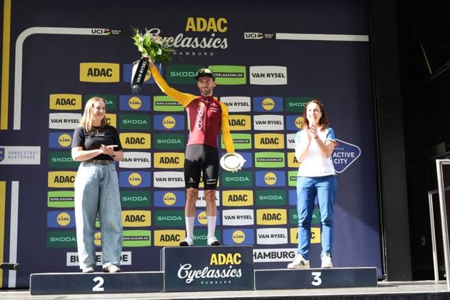 Benjamin Thomas stands on the top step of the podium at the ADAC Cyclassics Hamburg and raises the winner’s bouquet in celebration of his victory in the mountain classification; in his other hand, he holds the winner’s trophy. Two hostesses flank the podium, while sponsor logos from ADAC, Skoda, Lidl, and Van Rysel fill the background. Benjamin Thomas (147) celebrates at the awards ceremony for the ADAC Cyclassics Hamburg. Copyright: xspeedshotx xSarahxWolfx