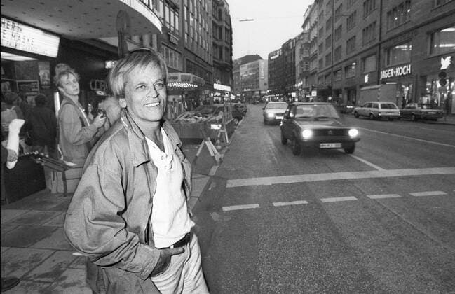 Klaus Kinski (Schauspieler) 10 85 vZ Klaus Kinski im Oktober 1985 in Hamburg Deutschland deutsch Deutscher Mann Schauspiel TV Fernsehen Film Kino Autor grauhaarig weisshaarig kurzhaarig leger Jacke Hemd quer halb stehend draußen lächelnd Stadt Innenstadt Straße Autos PKW fahrend sw