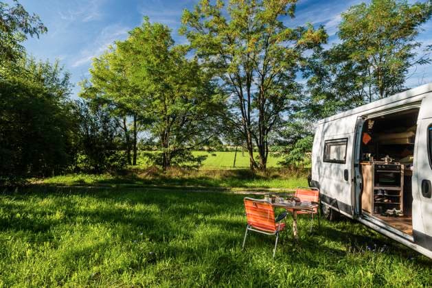 A camper stands between trees on a sunny summers day on a meadow in the countryside, a laid breakfast table with chairs stands in front of the sliding door of the camper Regensburg, Bavaria, Germany In the old town of Regensburg