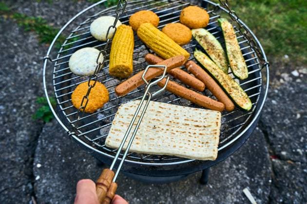 Bavaria, Germany - 26 July 2024: A colorful mix of vegetarian delicacies such as corn on the cob, vegan sausages, zucchini and grilled cheese sizzle on a charcoal grill. A man turns a meatless sausage with a pair of tongs