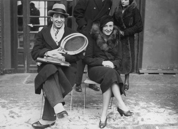 Orig. caption... RENE LACOSTE TURNS TO TENNIS AGAIN. Famous French star and international champion who figured so prominently in bringing the Davis Cup to France, but who has been in retirement, attends the Paris-London Inter-city tournament on covered courts with his equally famous wife, the former Mlle. Thion de la Chaume, golf champion. But RenÃ carried two tennis rackets and gave rise to speculation about the possibility of his taking part in the Davis Cup defense this year. It is known that he has been practicing regularly and every French fan hopes he will be in shape to join Henri Cochet and turn back any challenge. Lacoste and his wife are here seen on the sidelines in happy contemplation of the prospects of the game. Note R Lacost  Copyright: xx THE LACOSTE COUPLE