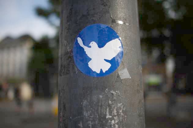 Flensburg, Schleswig Holstein A round blue sticker with a white dove of peace can be seen on a grey, slightly weathered lamppost The dove is depicted flying and symbolizes peace The background of the image is blurred, indicating a busy urban environment The image has a documentary feel a
