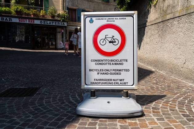 Limone sul Garda, Lombardy, Italy - August 7, 2025: Mobile traffic sign in Limone sul Garda: red circle with bicycle, note: bicycles only allowed with hand guidance. Pedestrians pass on the cobblestone square in the old town. Rule for the pedestrian zone
