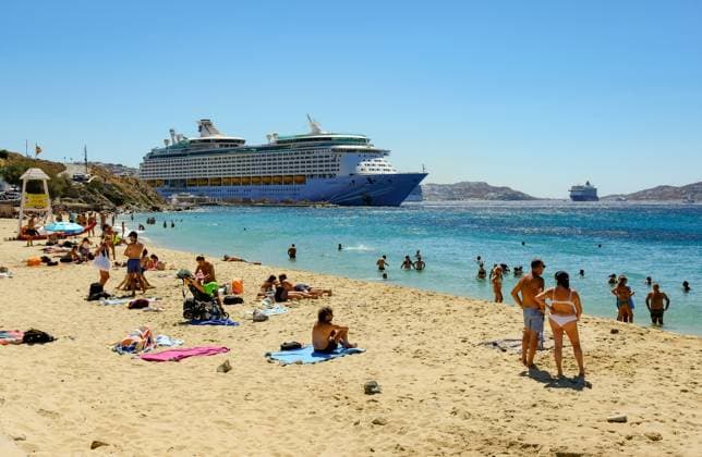 Mykonos, Cyclades, Greece Cruise ship Voyager of the Seas moored in Mykonos harbor Bay In front, tourists and locals bathe on the sandy beach Paralia Agios Stefanos Mykonos Mykonos Greece