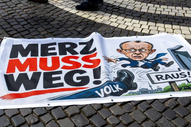 Augsburg, Bavaria, Germany May 16, 2026: Demonstration in Augsburg against the current federal government led by Chancellor Friedrich Merz. Under the slogan: Merz must go! Banner with the inscription: MERZ MUST GO! Featuring an illustration of a foot standing on the word PEOPLE and an illustration of Chancellor Friedrich Merz (CDU) flying through a door with a sign reading: GET OUT!