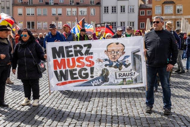 Augsburg, Bavaria, Germany May 16, 2026: Demonstration in Augsburg against the current federal government led by Chancellor Friedrich Merz. Under the slogan: Merz must go! Participants holding a banner reading: MERZ MUST GO! Featuring an illustration of a foot standing on the word PEOPLE and an illustration of Chancellor Friedrich Merz (CDU) flying through a door with a sign reading: GET OUT!