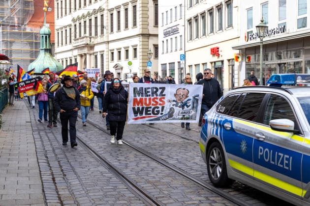 Augsburg, Bavaria, Germany May 16, 2026: Demonstration in Augsburg against the current federal government led by Chancellor Friedrich Merz. Under the slogan: Merz must go! Participants holding a banner reading: MERZ MUST GO! Featuring an illustration of a foot standing on the word PEOPLE and an illustration of Chancellor Friedrich Merz (CDU) flying through a door with a sign reading: GET OUT!
