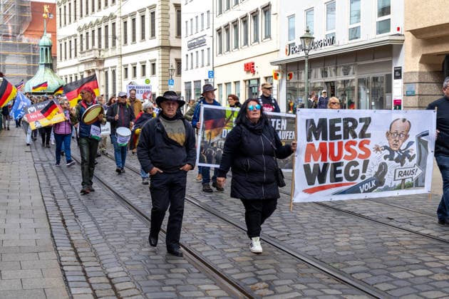 Augsburg, Bavaria, Germany May 16, 2026: Demonstration in Augsburg against the current federal government led by Chancellor Friedrich Merz. Under the slogan: Merz must go! Participants holding a banner reading: MERZ MUST GO! Featuring an illustration of a foot standing on the word PEOPLE and an illustration of Chancellor Friedrich Merz (CDU) flying through a door with a sign reading: GET OUT!