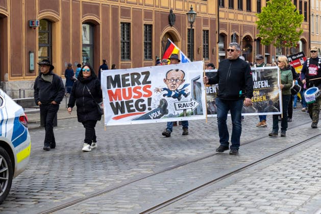 Augsburg, Bavaria, Germany May 16, 2026: Demonstration in Augsburg against the current federal government led by Chancellor Friedrich Merz. Under the slogan: Merz must go! Participants holding a banner reading: MERZ MUST GO! Featuring an illustration of a foot standing on the word PEOPLE and an illustration of Chancellor Friedrich Merz (CDU) flying through a door with a sign reading: GET OUT!