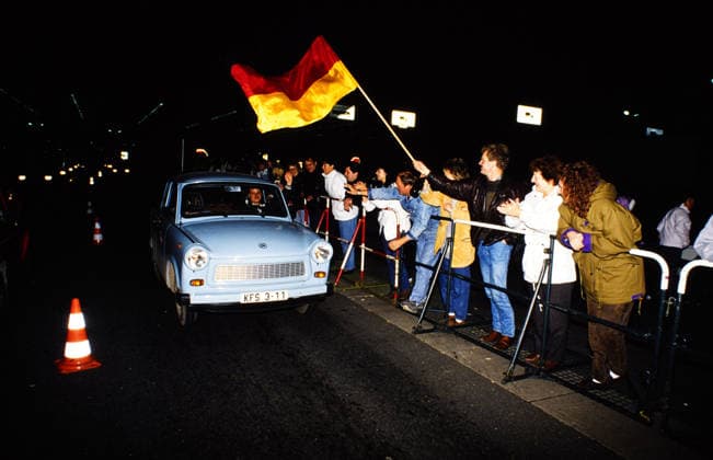 a light blue Trabant drives at night via the open Border, Jubelnde People with a German flag Standing on Roadside and welcome the Occupants, Germany, 1989 Berlin Berlin Germany Copyright: xUnitedxArchivesx xSvenxSimonxArchive SVS_94060000002