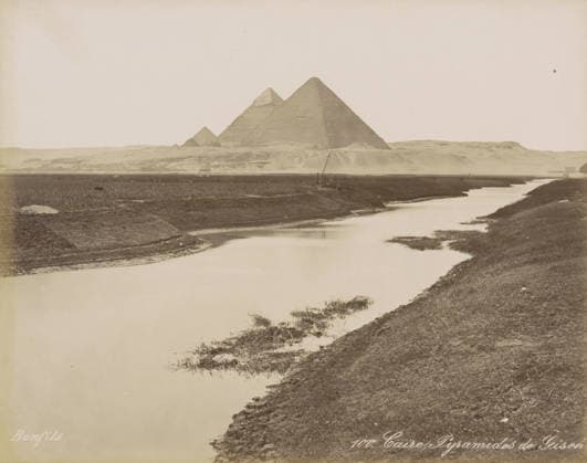 RECORD DATE NOT STATED Caire, Pyramids of Giza, View of the Pyramids of Giza near Cairo, photograph, Part of Travel Album with 80 Photos of Egypt., negative number: 100 , photographer: Maison Bonfils, Giza, c. 1870 - c. 1898, North Africa, pasteboard, paper, albumen print, height 220 mm x width 282 mm, pyramid (historical grave form), Ottoman Empire, Cairo (city), A wide sepia-toned landscape showing a calm winding river in the foreground with grassy and muddy banks, a low flat plain beyond the river, and three large pyramidal stone structures on a distant rise against a pale sky, with sparse vegetation and a small boat or wooden structure near the far bank. Copyright Penta Springs LCK260531_03313