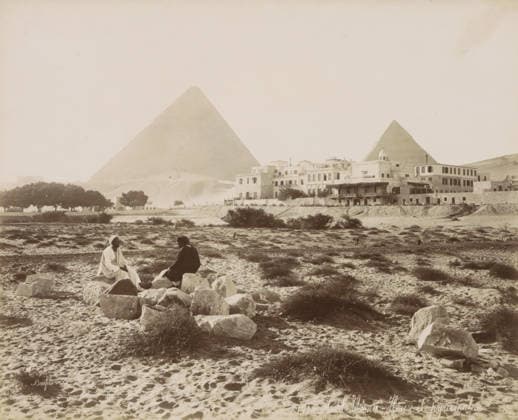 RECORD DATE NOT STATED Hotel Mena - House and pyramids, View of the Pyramids of Giza with Hotel Mena House in the foreground, photograph, Part of Travel Album with 80 Photos of Egypt., negative number: 1434 , photographer: Maison Bonfils, Giza, c. 1886 - c. 1898, North Africa, pasteboard, paper, albumen print, height 221 mm x width 272 mm, hotel, hostelry, inn, pyramid (historical grave form), A wide sandy landscape with scattered low shrubs and rocks in the foreground shows two seated figures on a cluster of stones, one wearing a light robe and head covering and the other in darker clothing and a brimmed hat, facing each other. Behind them a built cluster of multi level stone and stucco buildings with flat roofs and a small domed structure runs along a raised Copyright Penta Springs LCK260531_03314
