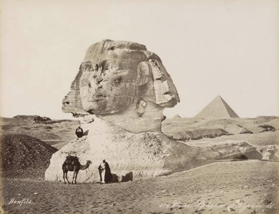 RECORD DATE NOT STATED Sphinx and pyramids of Giza, Caire, Sphynx et Pyramide, photograph, Part of Travel album with 80 photos of Egypt., negative number: 111 , photographer: Maison Bonfils, Gizeh, c. 1870 - c. 1898, North Africa, Giza, pasteboard, paper, albumen print, height 220 mm x width 282 mm, pyramid (historical grave form), piece of sculpture, reproduction of a piece of sculpture (sculpture), A large weathered stone sculpture of a human head and neck dominates the foreground, showing layered textures and erosion lines on its surface. A person sits on the stone near the sculpture™s base, and another person stands in front of the base beside a camel, which has a saddle and some coverings. A small figure is positioned on a ledge on the sculpture™s neck. Copyright Penta Springs LCK260531_03315