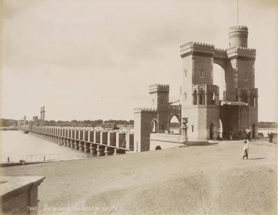 RECORD DATE NOT STATED View of a dam on the Nile near Cairo, General view of the Nile dam, photograph, Part of a travel album with 80 photos of Egypt., check stamp: 1445 , photographer: Maison Bonfils, Egypt, c. 1870 - c. 1898, North Africa, pasteboard, paper, albumen print, height 221 mm x width 281 mm, dam, Ottoman Empire, Cairo (city), A wide riverside scene shows a long bridge extending into the distance with evenly spaced vertical supports rising from the water, leading to a series of brick towers with crenellated tops and arched openings; a larger ornate brick gateway with rounded turrets and decorative arches stands near the foreground, showing carved stonework and columned openings, while people in period clothing walk and stand near the entrance and on Copyright Penta Springs LCK260531_03316