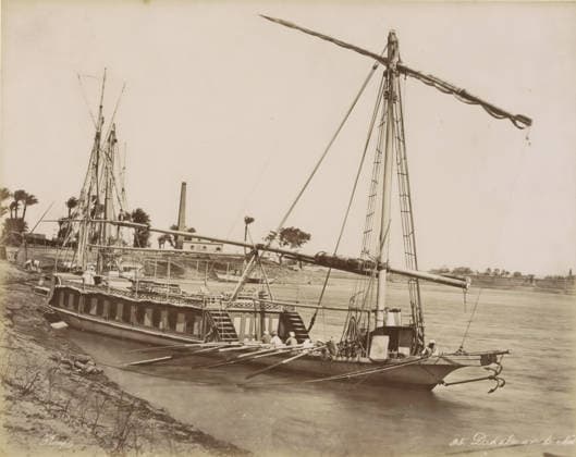 RECORD DATE NOT STATED Dahabie on the Nile, Passenger boat (dahabie) on the Nile, photograph, Part of Travel Album with 80 Photos of Egypt., negative number: 95 , photographer: Maison Bonfils, Egypt, c. 1870 - c. 1898, North Africa, pasteboard, paper, albumen print, height 222 mm x width 278 mm, sailing-ship, sailing-boat, Nile, A large boat with tall masts and long spars is docked along a riverbank, showing a wooden hull with a row of cabin windows and an open deck with railings and steps leading down; several ropes, rigging lines, and ladders run up the masts, and a group of people wearing light clothing and head coverings sit or stand near the stern and along the deck; the river water is calm and stretches across the image with another boat partly visible in Copyright Penta Springs LCK260531_03317