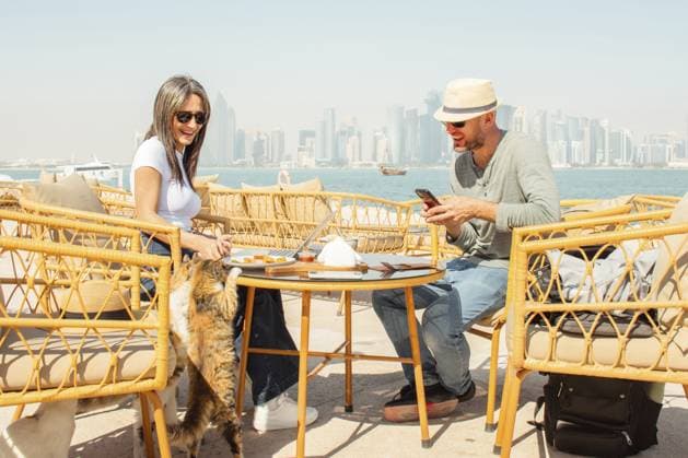 Nahaufnahme Mann und Frau sitzen im Café Doha Corniche im Freien. Frau spielt beim Füttern von Katzen, Mann mit Smartphone hat Spaß. Korbstühle, Tisch, Essen, Kaffee, Laptop, Taschen. Aussichtspunkt auf die Skyline von West Bay Close up Man and woman sitting in outdoor cafe Doha Corniche. Woman playing feeding cats, man using smartphone having fun. Wicker chairs, table, food, coffee, laptop, bags. West Bay skyline viewpoint Copyright: imageBROKER EvaldasxMikoliunas ibxemi18090526.jpg ,model released, Symbolfoto ,property released