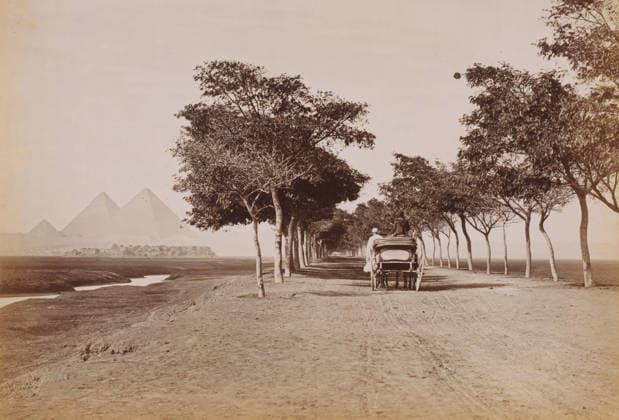 RECORD DATE NOT STATED Pyramid of Cheops, Grande pyramide de Chà ops, photograph, number: 113 , photographer: Fà lix Bonfils, Giza, 1867 - 1885, North Africa, pasteboard, paper, albumen print, height 200 mm x width 279 mm, height 396 mm x width 290 mm, pyramid (historical grave form), A long straight dirt road lined with two rows of tall leafy trees forming a shaded avenue, a horse-drawn carriage with a covered back facing away from the viewer and two figures seated on and beside it, a narrow winding water channel running parallel to the road on the left, a cluster of low buildings beyond the channel, and three large pyramidal stone structures rising in the distant hazy background. Copyright Penta Springs LCK260531_18458