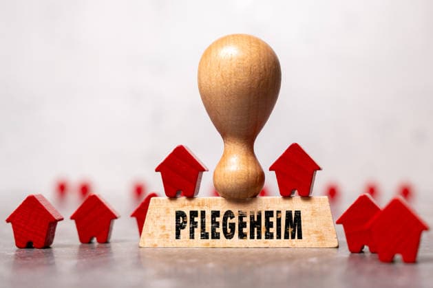 Bavaria, Germany - July 8, 2026: PHOTOMONTAGE. A wooden stamp bearing the inscription Nursing Home among small wooden houses symbolizes inpatient care and senior housing. The image represents nursing home beds, care services, demographic change, and long-term care policy.